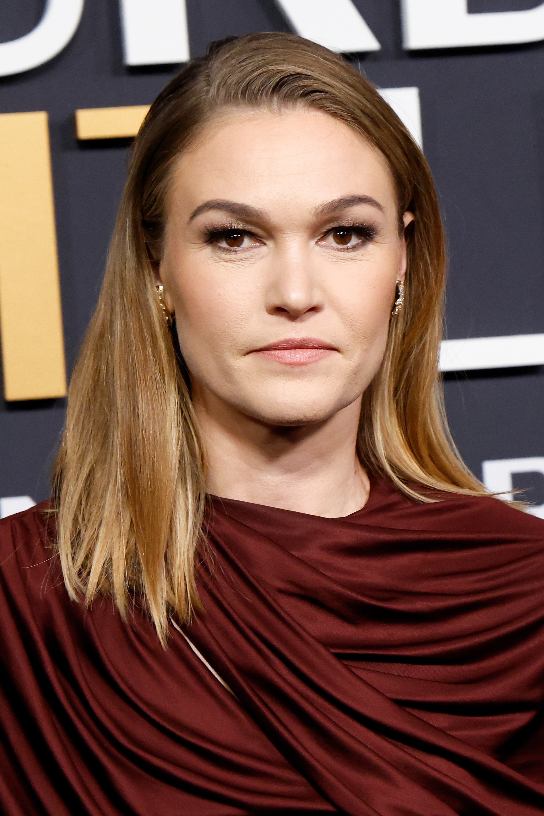 A person with shoulder-length hair wears an elegant draped gown at a formal event, standing in front of a patterned backdrop