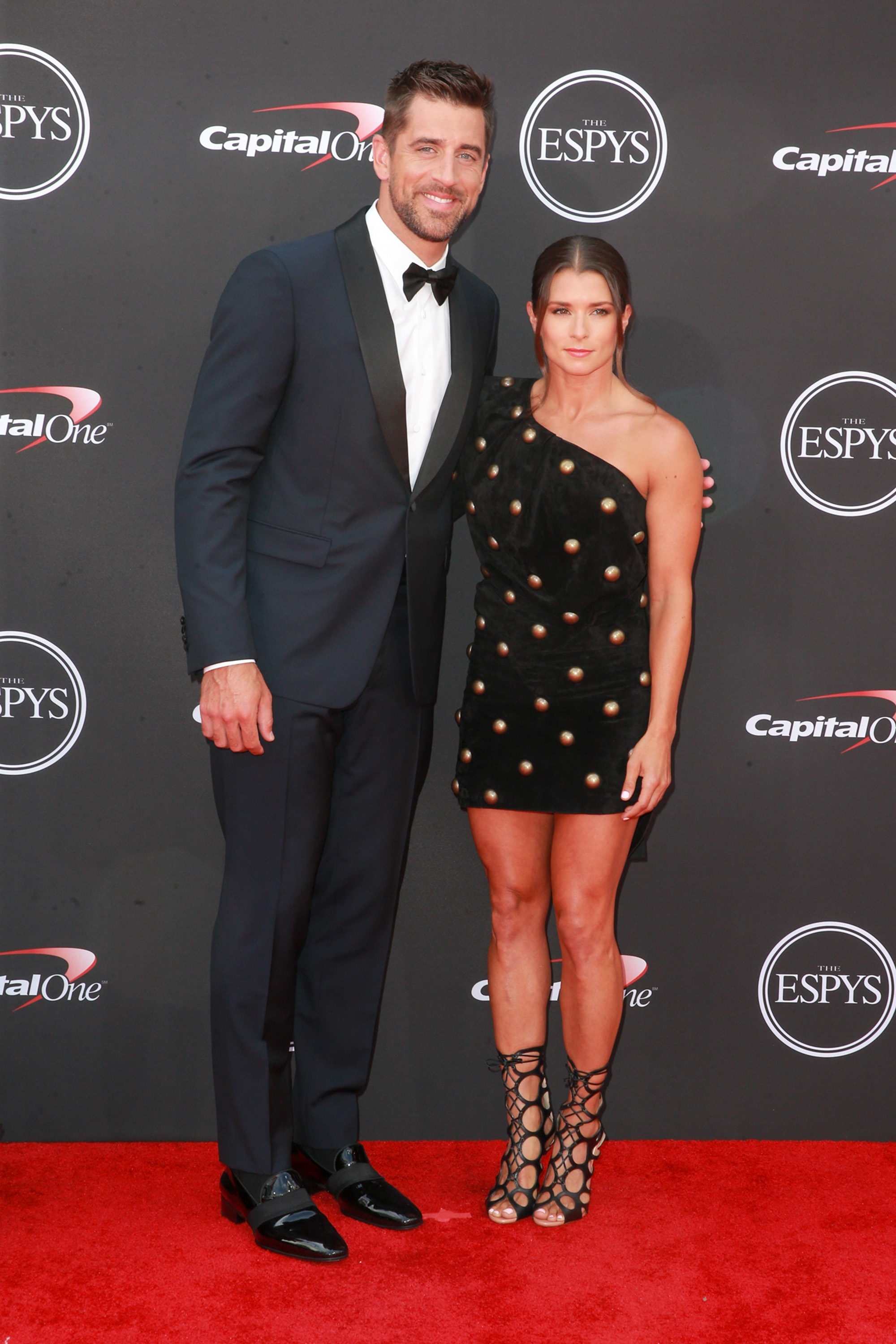 Aaron Rodgers in a tuxedo and Danica Patrick in a studded one-shoulder dress pose on the red carpet at an event