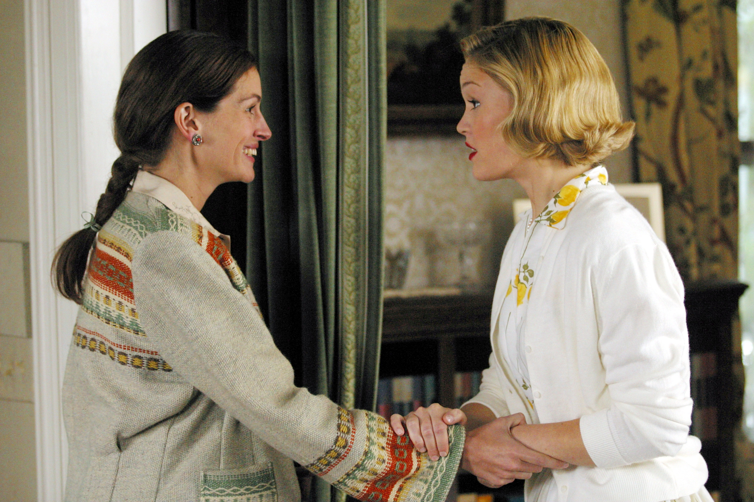 Two women in a room, one with intricate patterned clothing, shake hands while smiling, appearing engaged in conversation