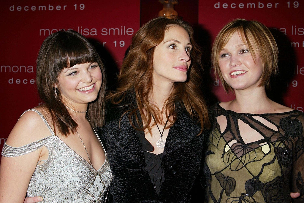 Three women smiling together at a film event. One wears a sparkly dress, another a black patterned dress, and the third a textured coat