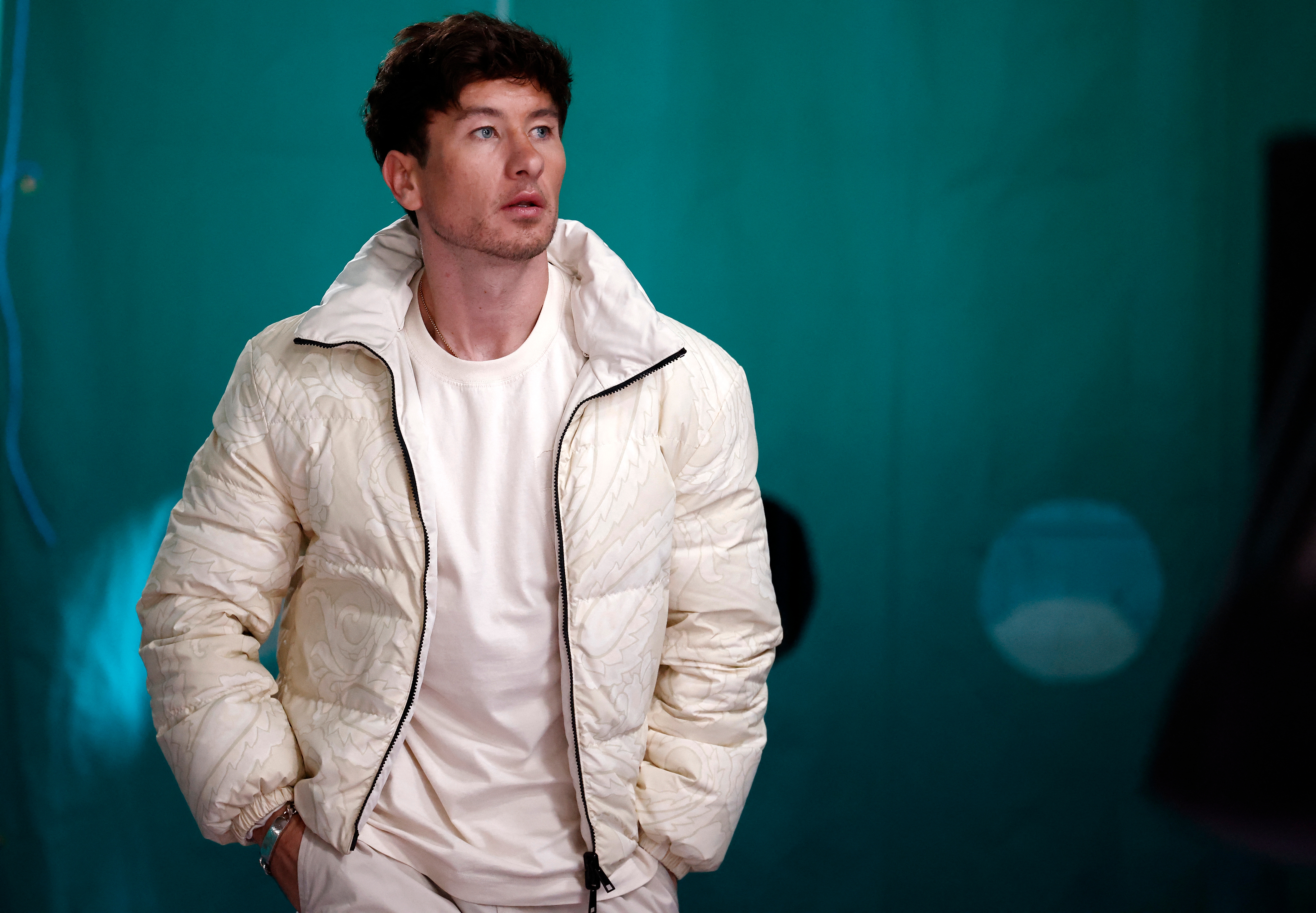 Barry Keoghan in a casual white quilted jacket and t-shirt, standing with hands in pockets. Blue-green background