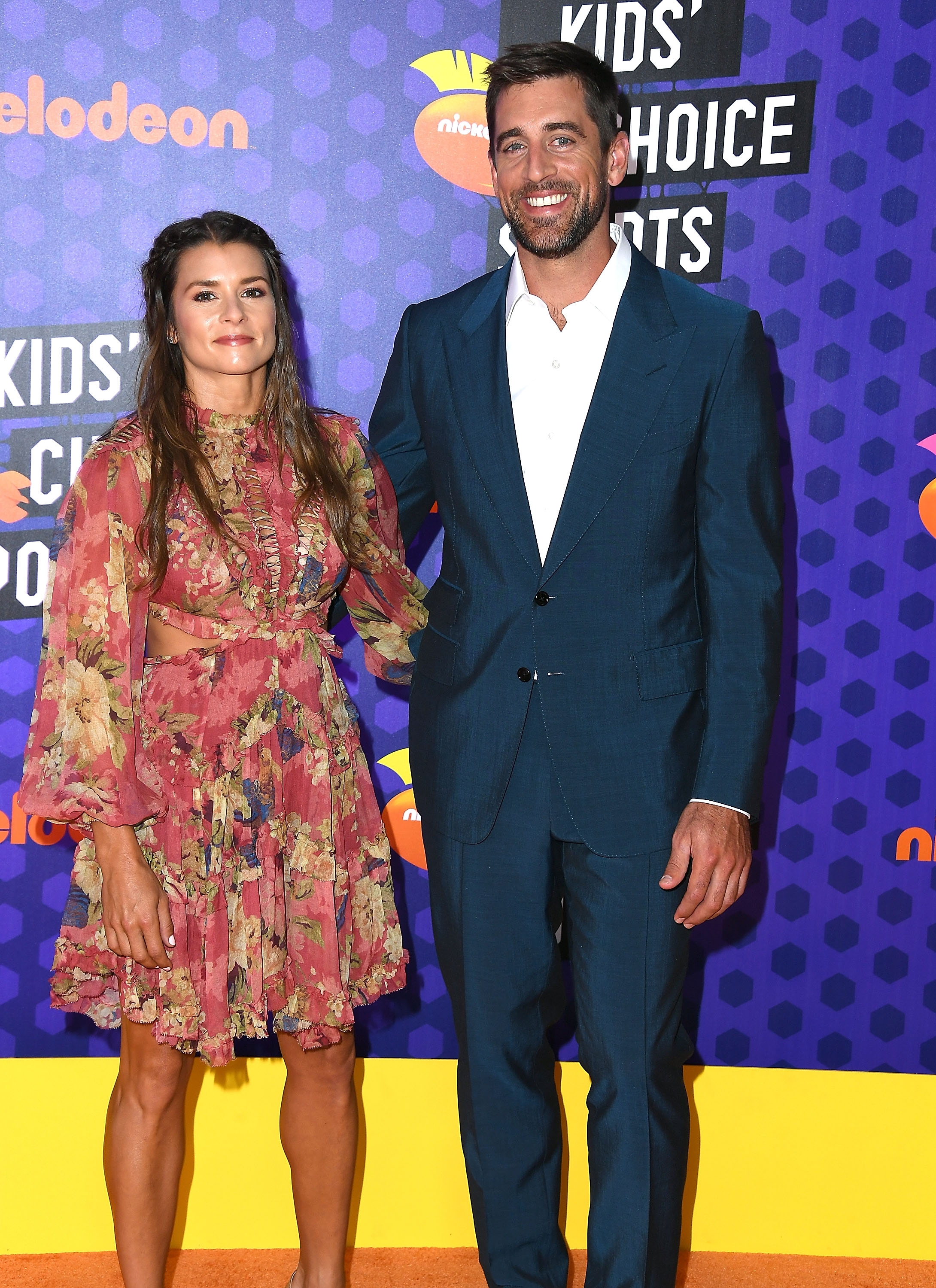 Danica Patrick and Aaron Rodgers on a red carpet; one in a floral dress, the other in a dark suit