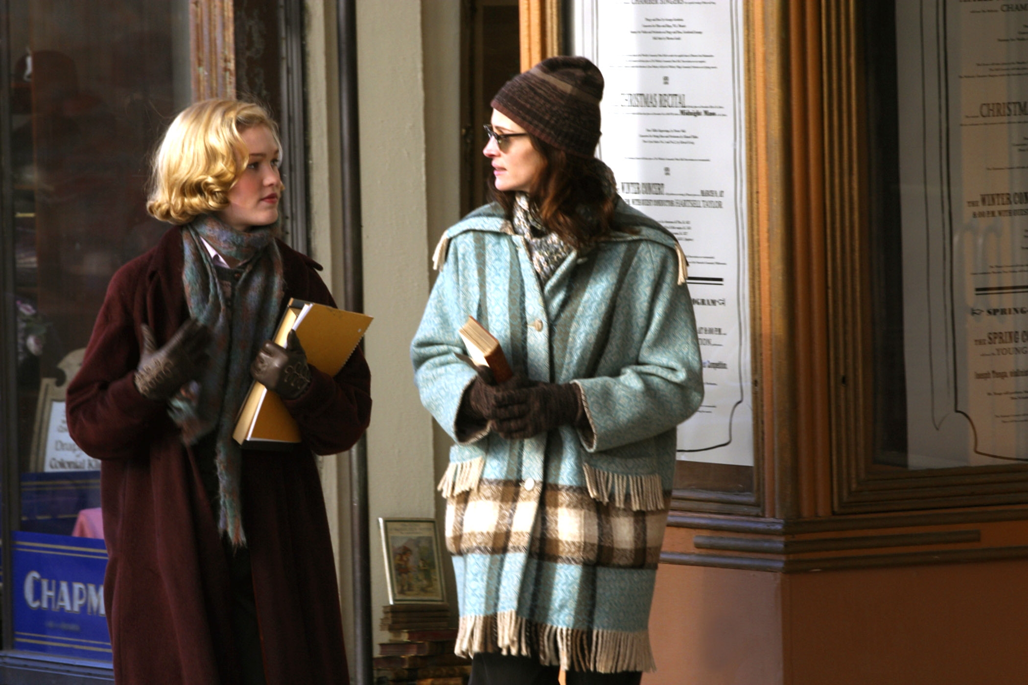 Two individuals in coats and scarves walk, holding books, in front of a storefront with posters. The scene suggests a casual, cool-weather day