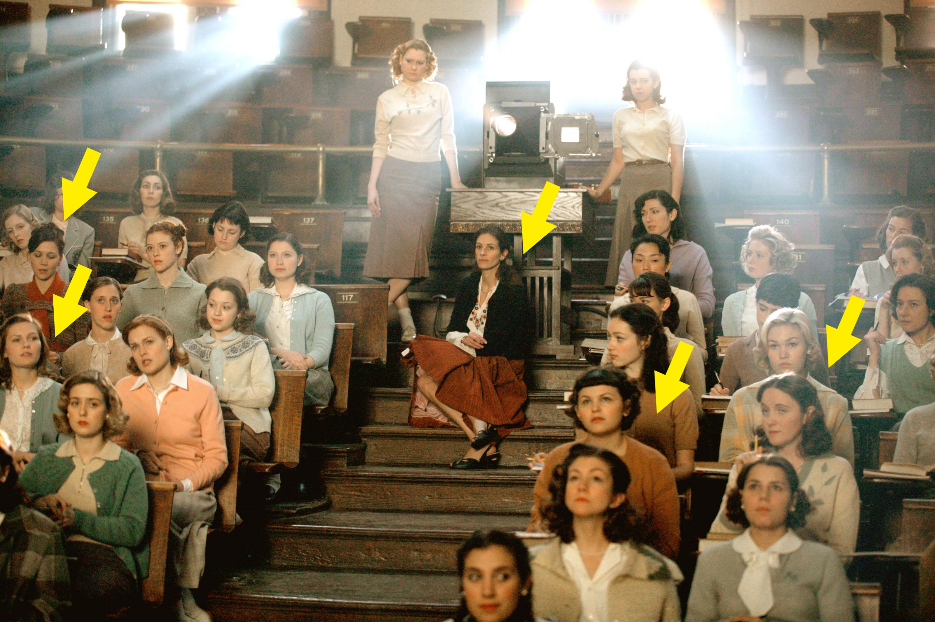 Women attending a lecture in a vintage classroom setting, some seated and others standing near a projector, all in mid-century attire