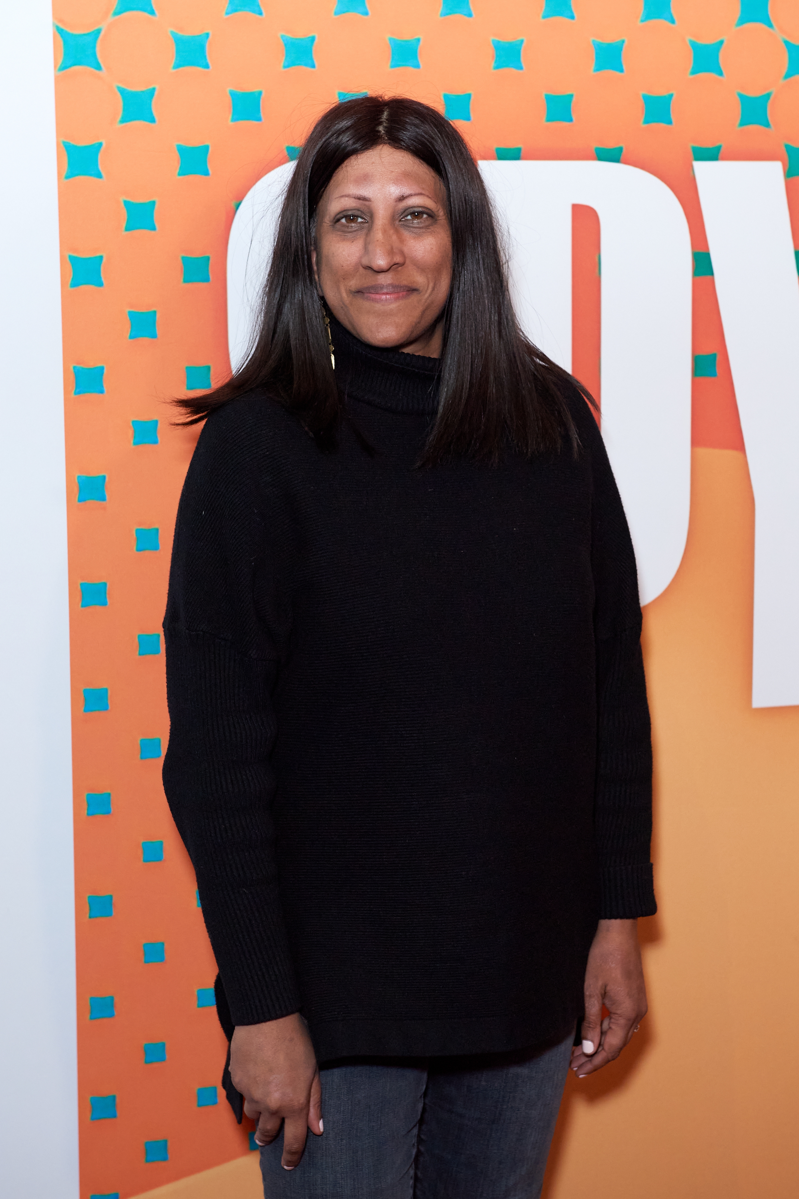 Person wearing a black turtleneck sweater and jeans, standing against a patterned backdrop at a TV event