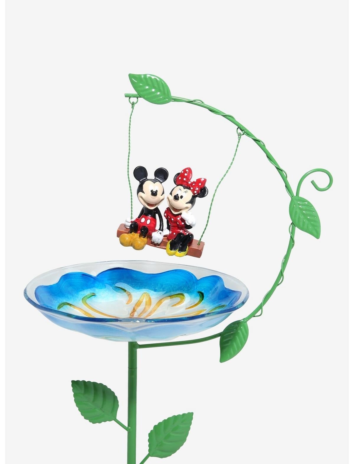 Mickey and Minnie Mouse figurines sit on a swing above a decorative glass birdbath with a floral design on a leafy stand