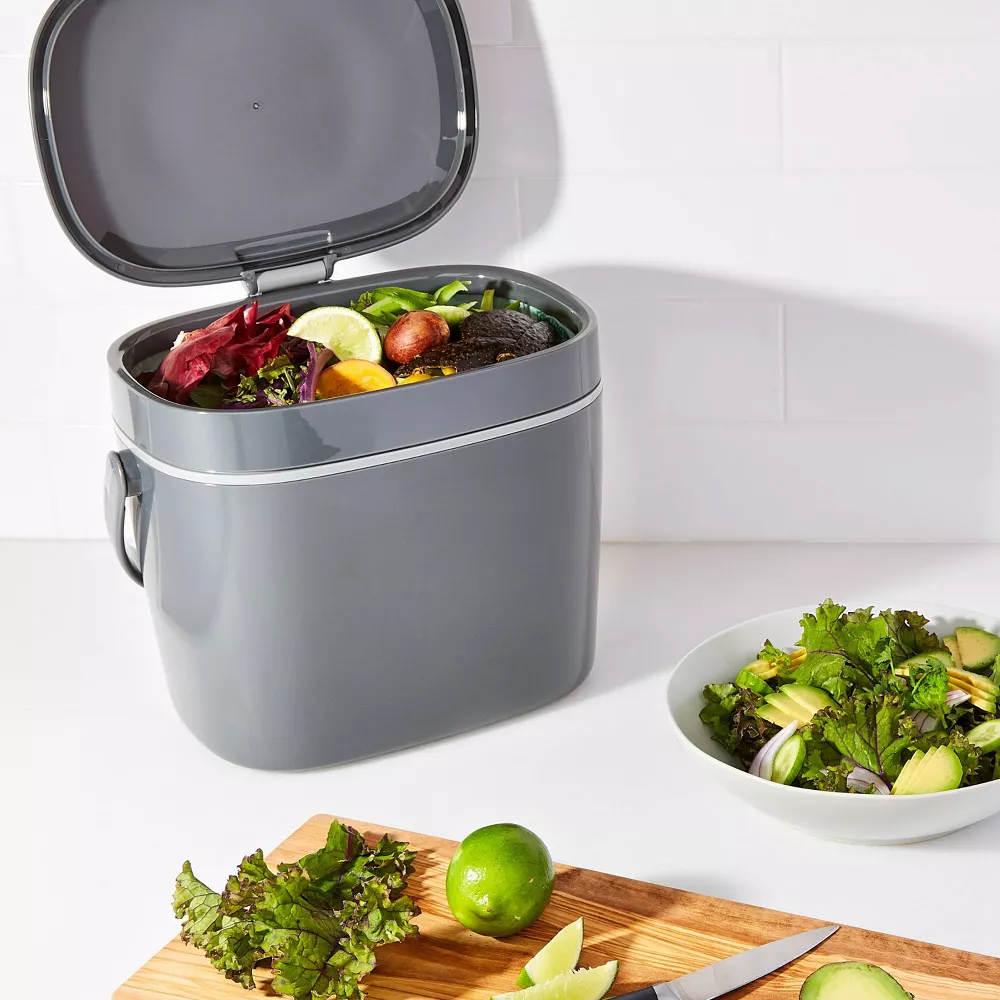 Countertop compost bin filled with food scraps, beside a salad and a cutting board with fresh vegetables and a knife