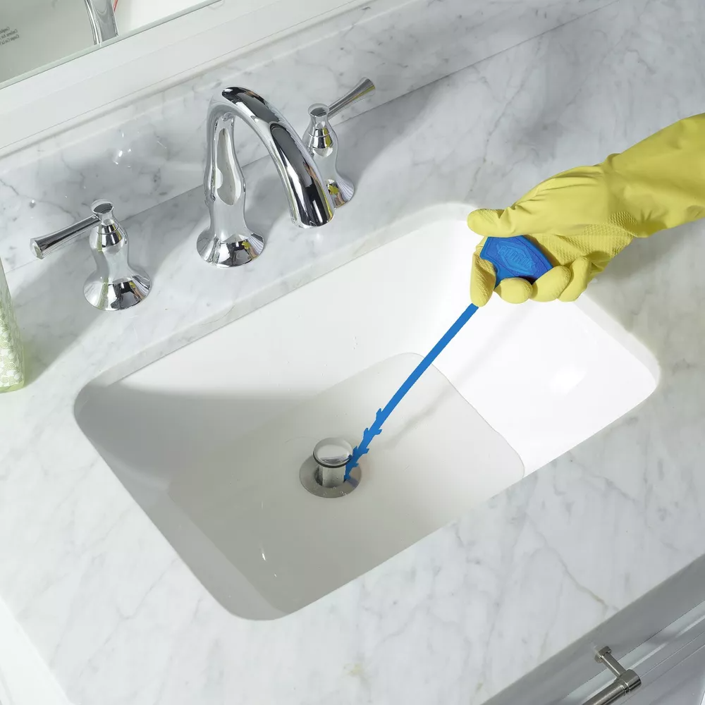 Person using a blue drain snake to unclog a sink, wearing yellow rubber gloves, suggesting a cleaning product being demonstrated for shopping purposes
