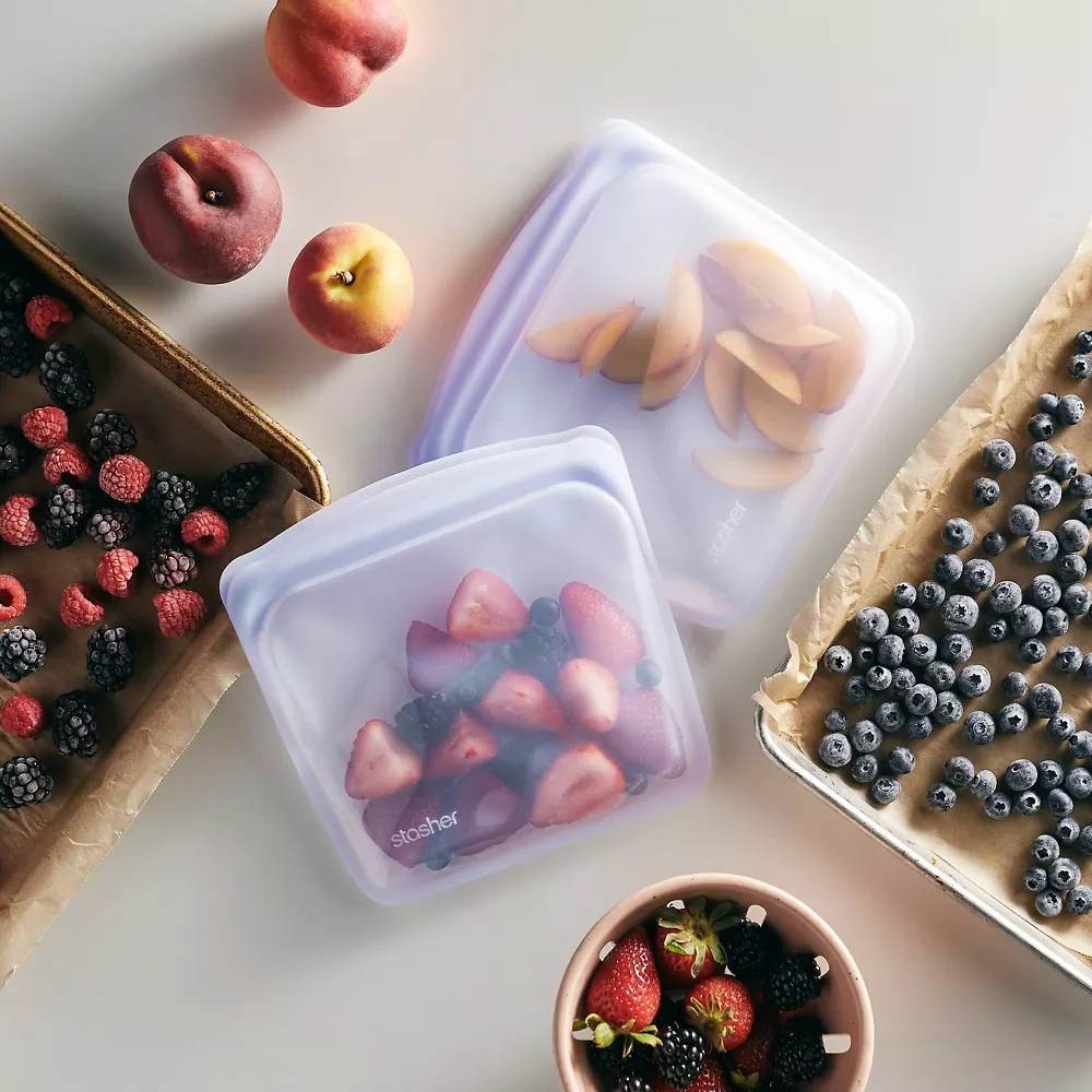 Reusable silicone bags filled with peach slices and strawberries, surrounded by trays of various fruits, promoting eco-friendly food storage options