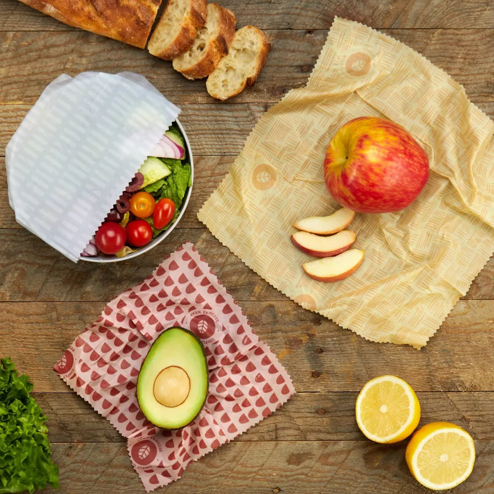 Reusable beeswax wraps covering salad, avocado, apple, lemon halves, and bread on a wooden table. Eco-friendly food storage option
