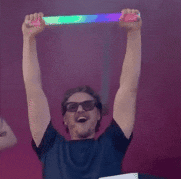 Person celebrates joyfully, holding a shiny rectangular object overhead, wearing sunglasses and a casual outfit