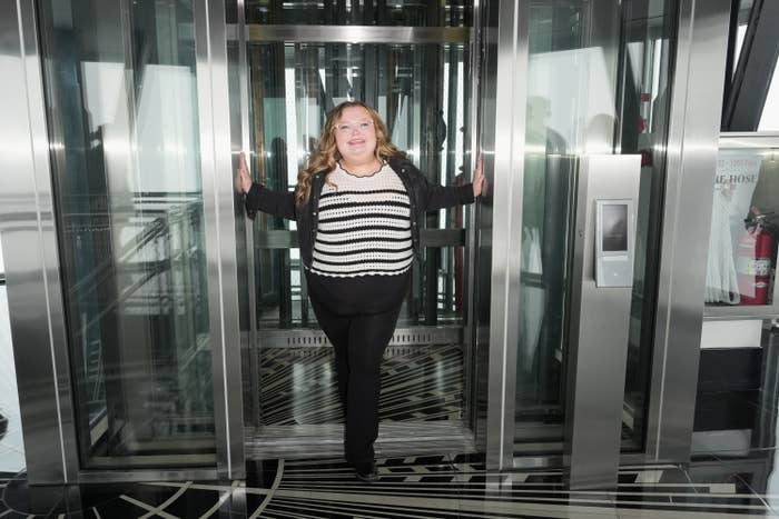Alana Thompson smiling in an elevator, wearing a striped top and black pants, standing confidently with hands on open doors