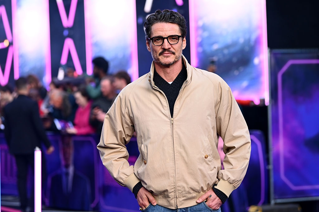Person in a casual beige jacket and jeans poses confidently on a bustling event carpet, with a crowd and vibrant background