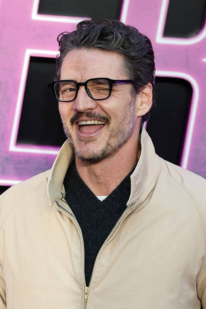 Man with glasses and short hair smiling playfully, wearing a casual jacket over a dark shirt, standing in front of a purple-lit backdrop