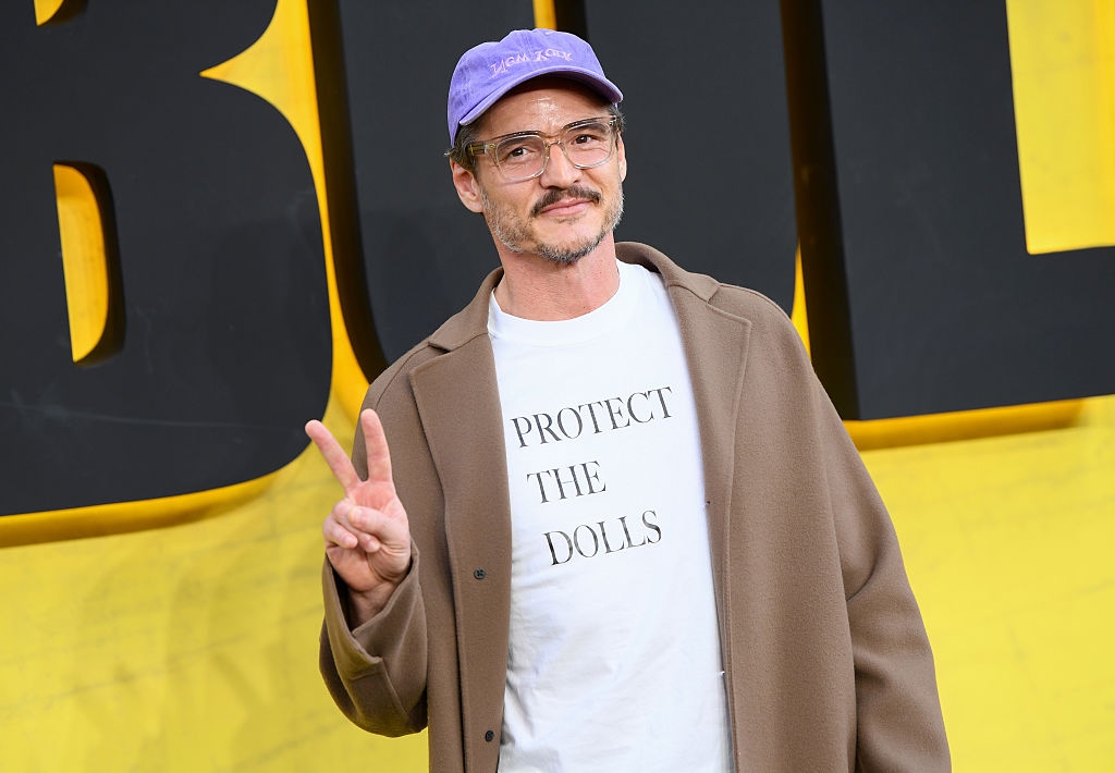 Person on a red carpet wearing a casual cap, glasses, white t-shirt with "PROTECT THE DOLLS" text, and a stylish coat, making a peace sign