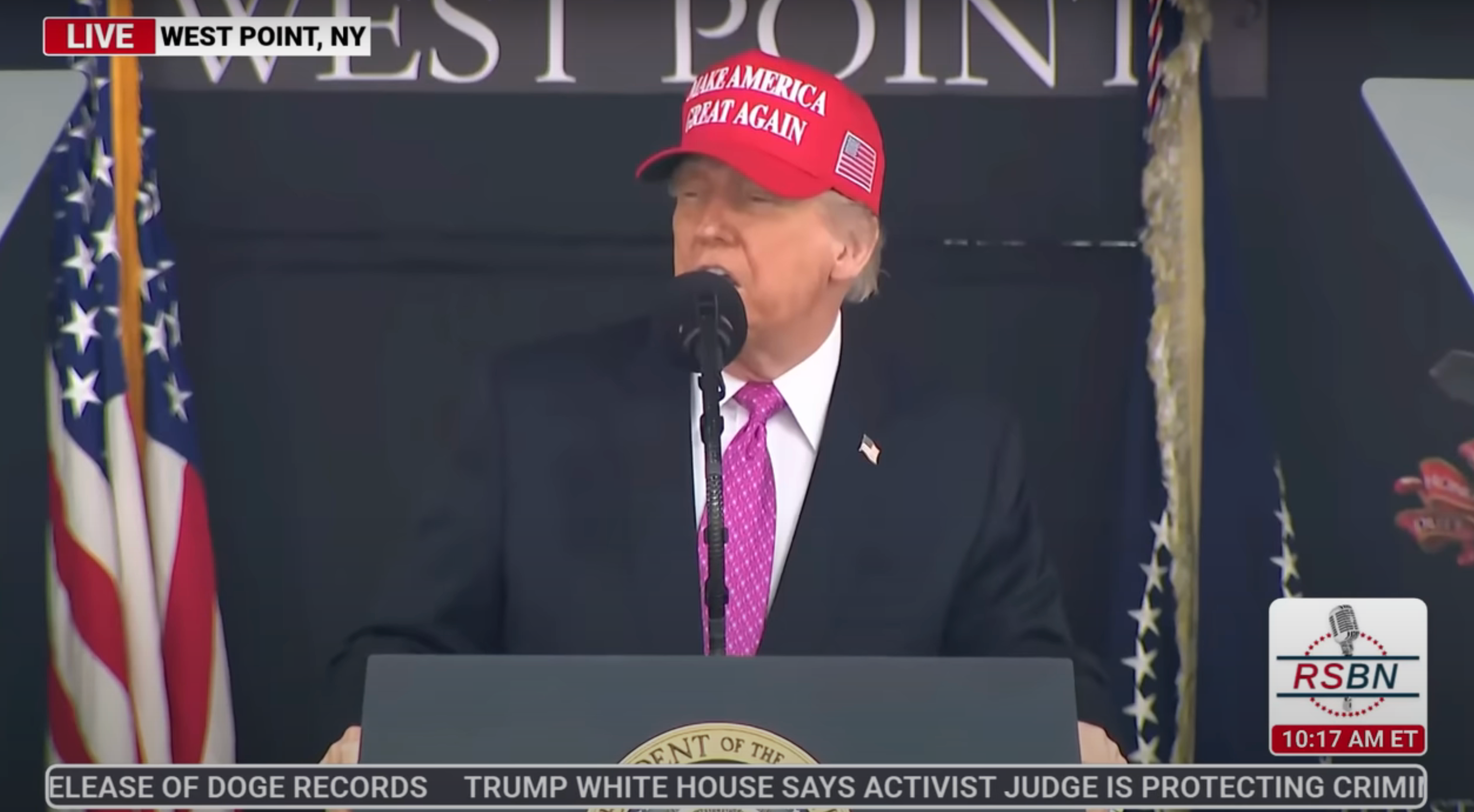 Person in a red hat and suit speaks at a podium with flags behind, during a live event in West Point, NY, as shown on a news broadcast