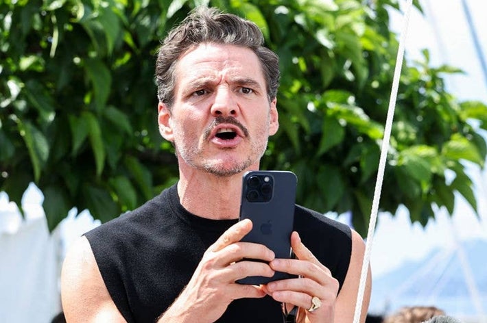A person outdoors holding a smartphone, appearing to speak or shout, wearing a sleeveless top with trees in the background