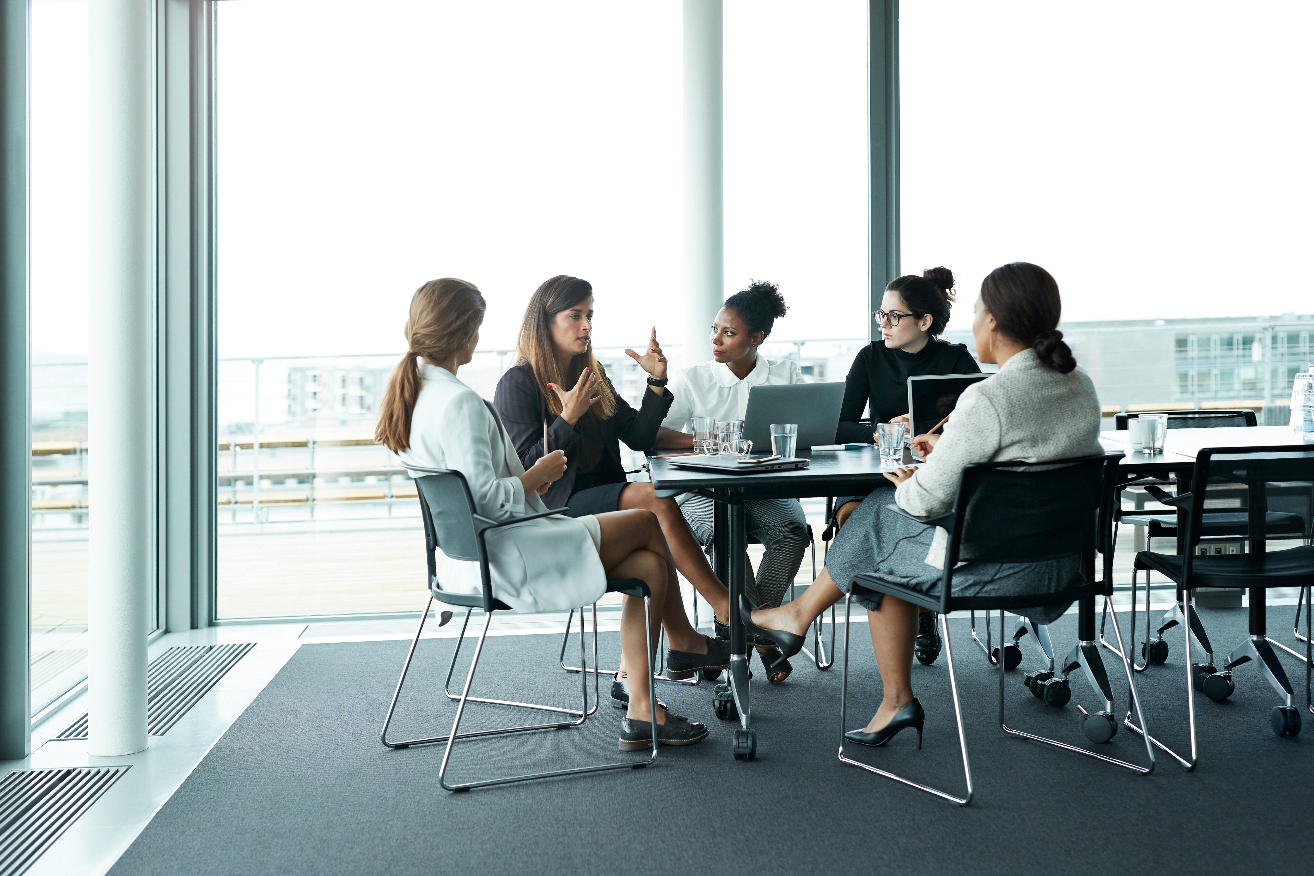 Group of five women in a modern office discussing around a table, with laptops and notebooks. They're engaged and interactive