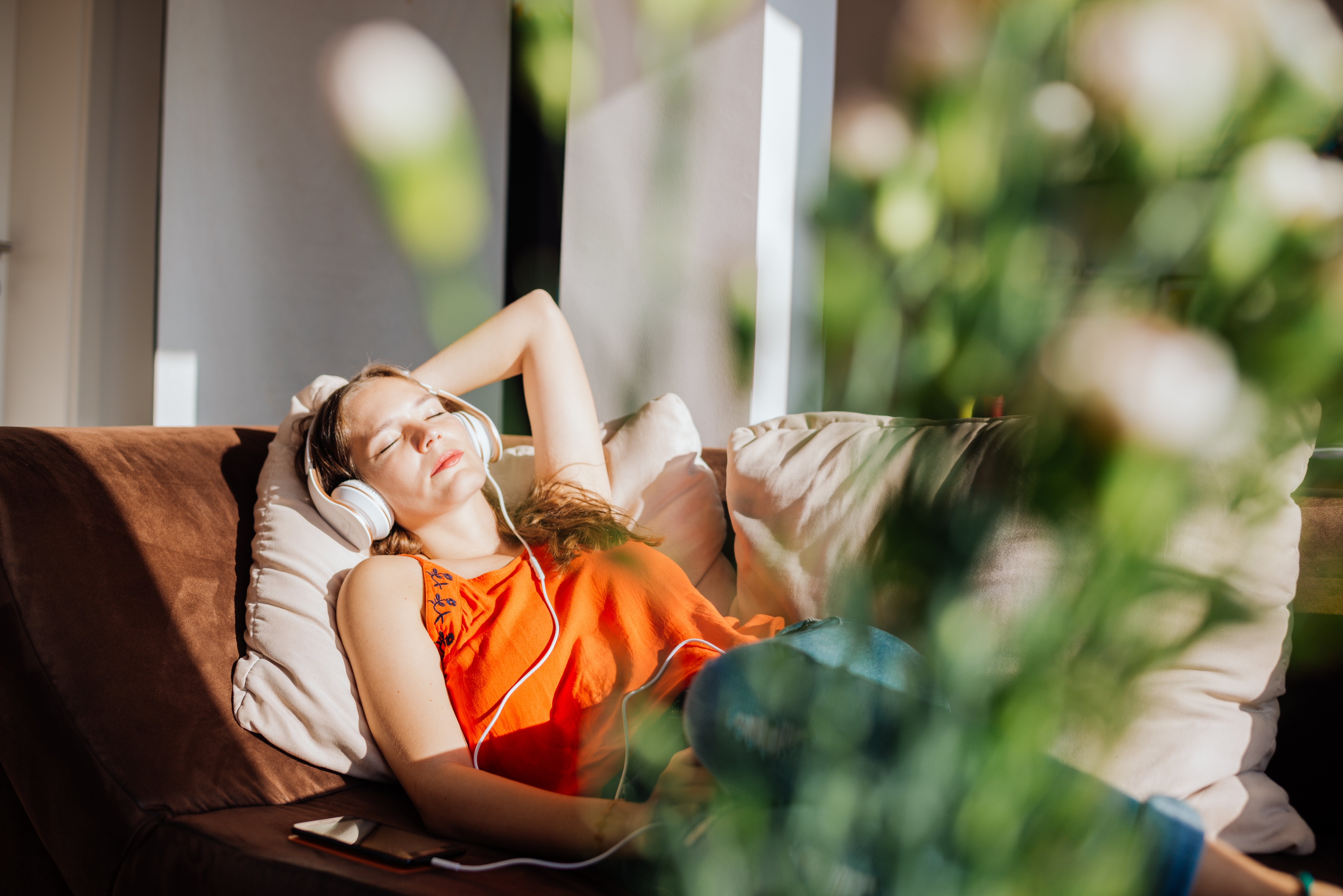 A woman relaxes on a couch with headphones on, eyes closed and arm resting on her head, enjoying a peaceful moment surrounded by blurred plants
