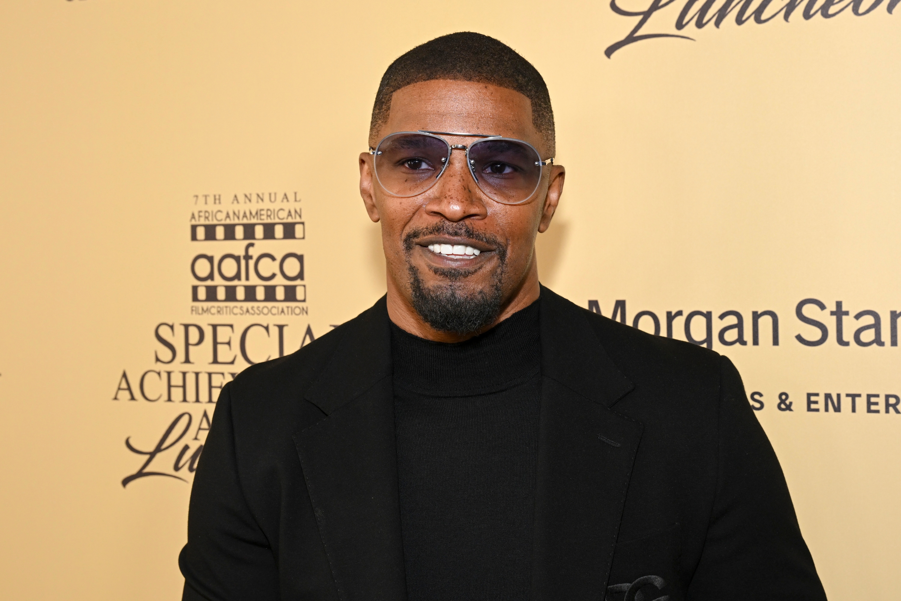 Jamie Foxx in a black suit and turtleneck smiles at an event. He wears sunglasses and has a trimmed beard