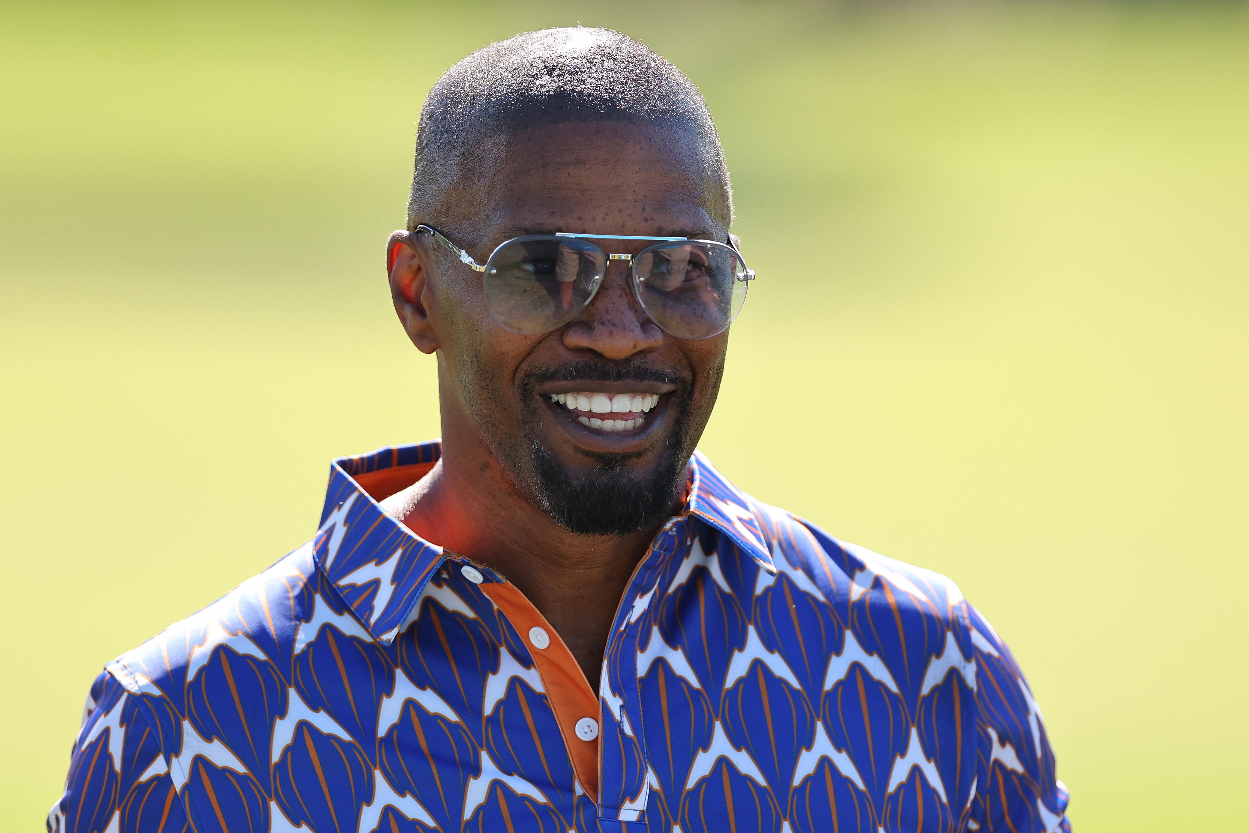 Jamie Foxx smiles while wearing aviator sunglasses and a vibrant patterned shirt with a pointed collar, in an outdoor setting