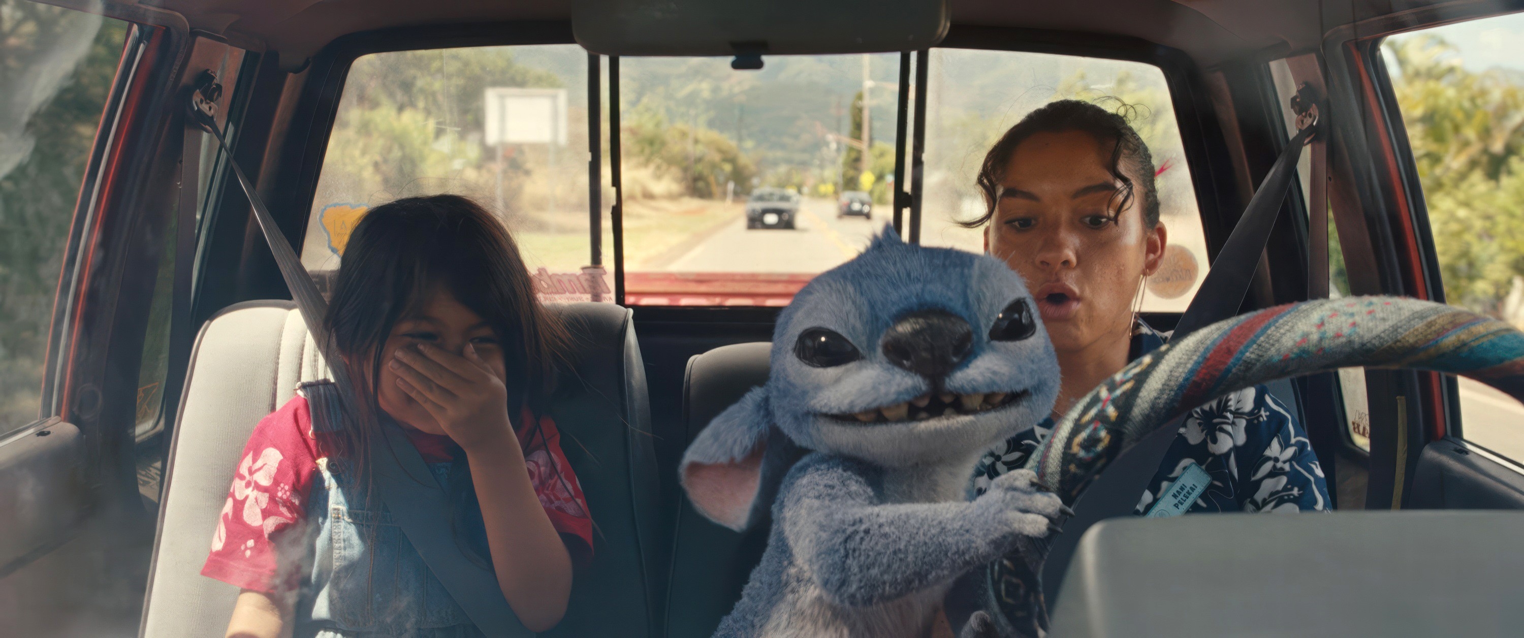 A woman drives a car on a road with a child laughing in the back seat and a blue animated creature in the front