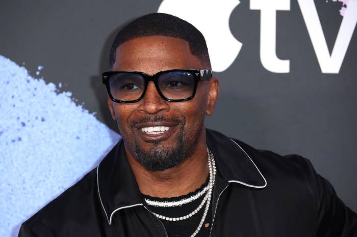 Jamie Foxx smiling at a media event, wearing glasses and a stylish black jacket with white trim and multiple necklaces