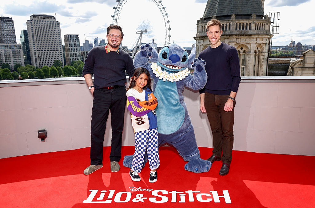 Three people pose with a Stitch character on a "Lilo & Stitch" red carpet with a cityscape and Ferris wheel in the background
