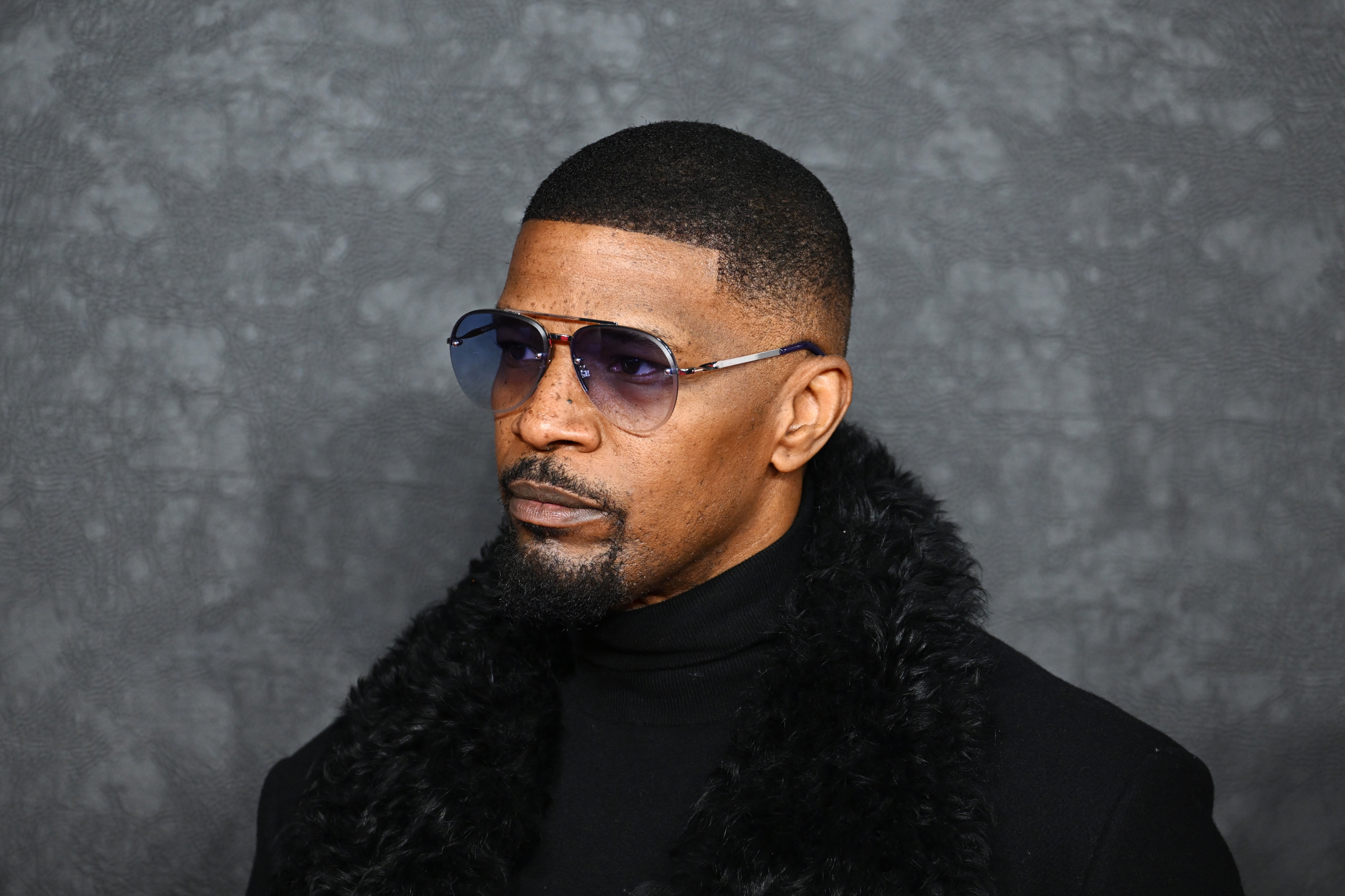 Jamie Foxx in sunglasses and black fur-trimmed coat, looking to the side, with a neutral expression against a textured background