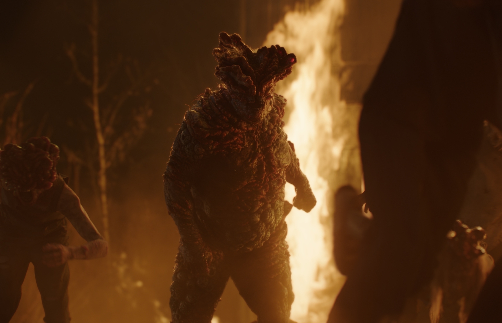 Una criatura monstruosa camina frente a un incendio en un entorno oscuro y ominoso, acompañada de otras figuras