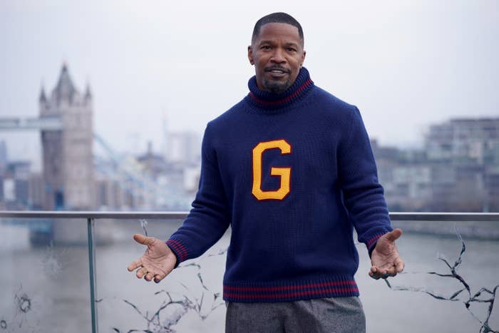 Jamie Foxx poses outdoors wearing a sweater with a large "G" on it, standing in front of a misty cityscape with a bridge in the background