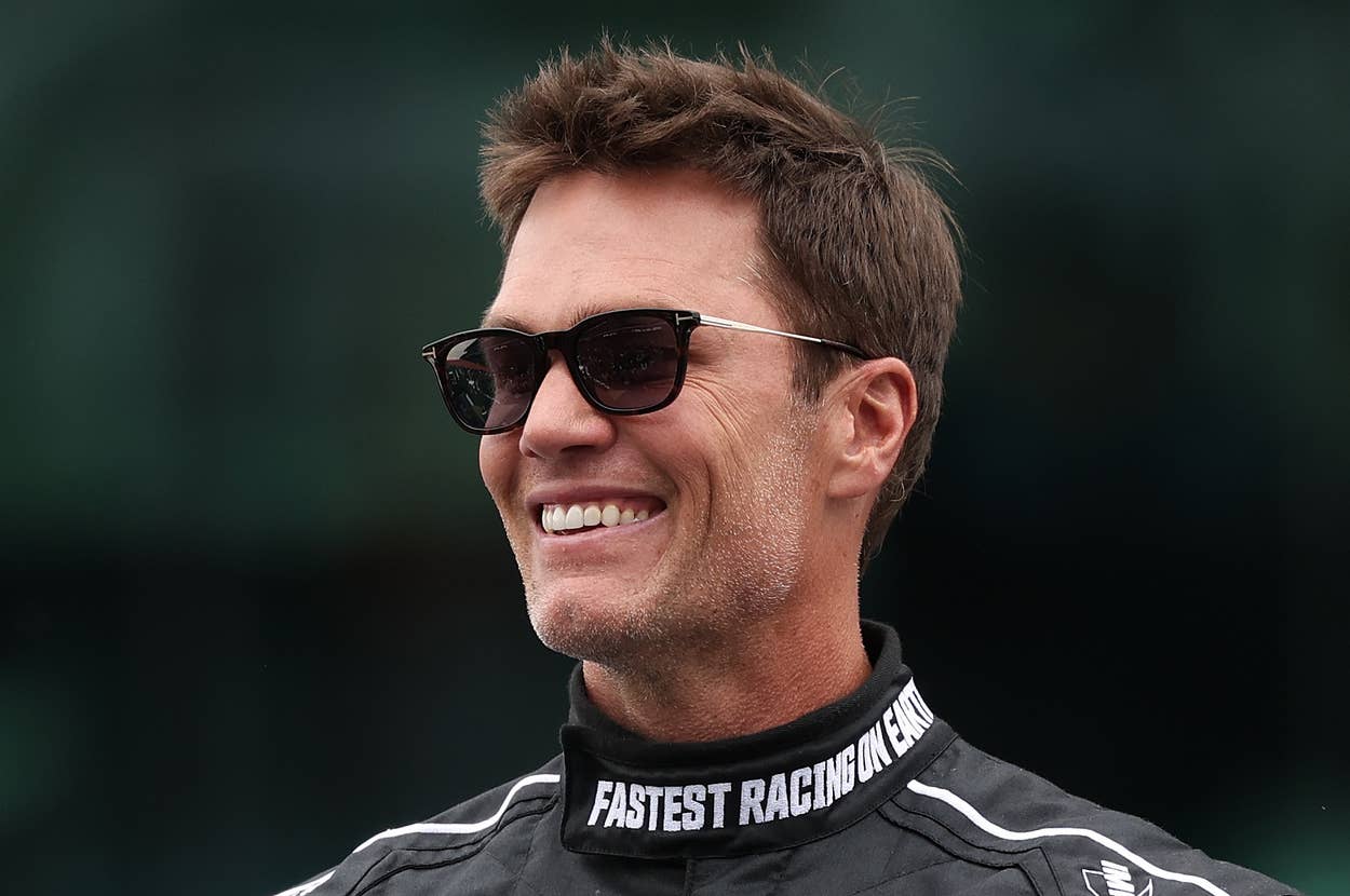Tom Brady in racing suit with sponsor logos, smiling and wearing sunglasses. They stand outdoors, looking to the side