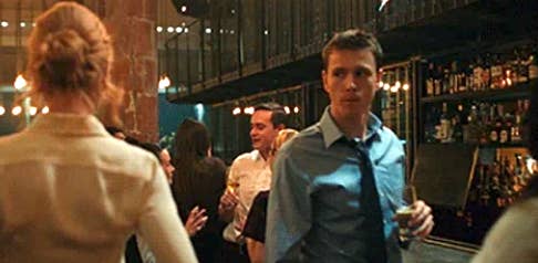 People socializing at a bar; a man in a blue shirt and tie holds a drink while a woman in a blouse stands nearby