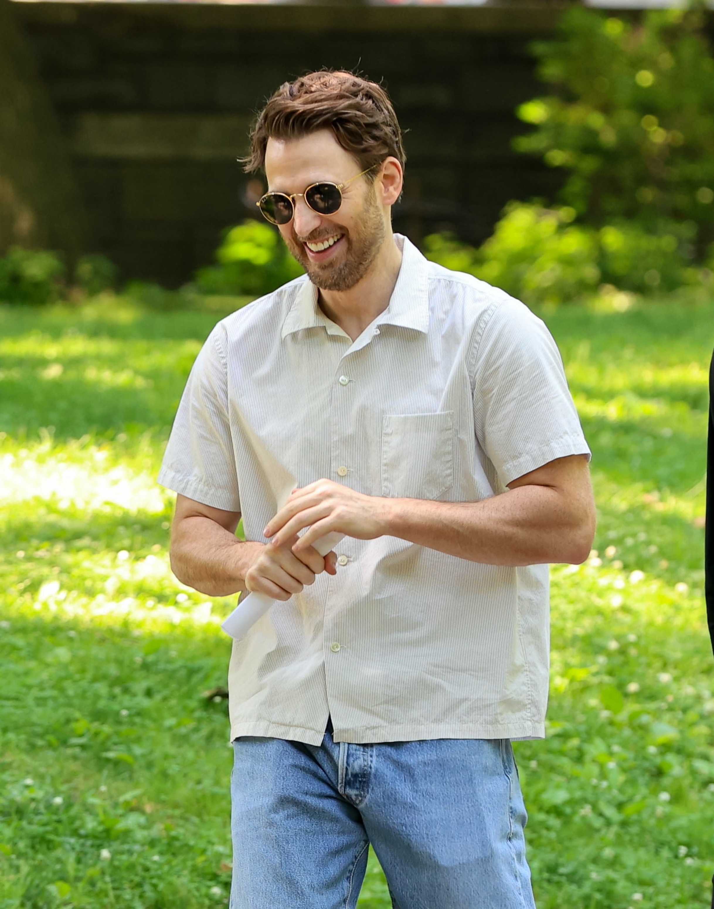 Chris Evans outdoors wearing a casual short-sleeve shirt and jeans, smiling while holding a rolled paper, in a relaxed setting