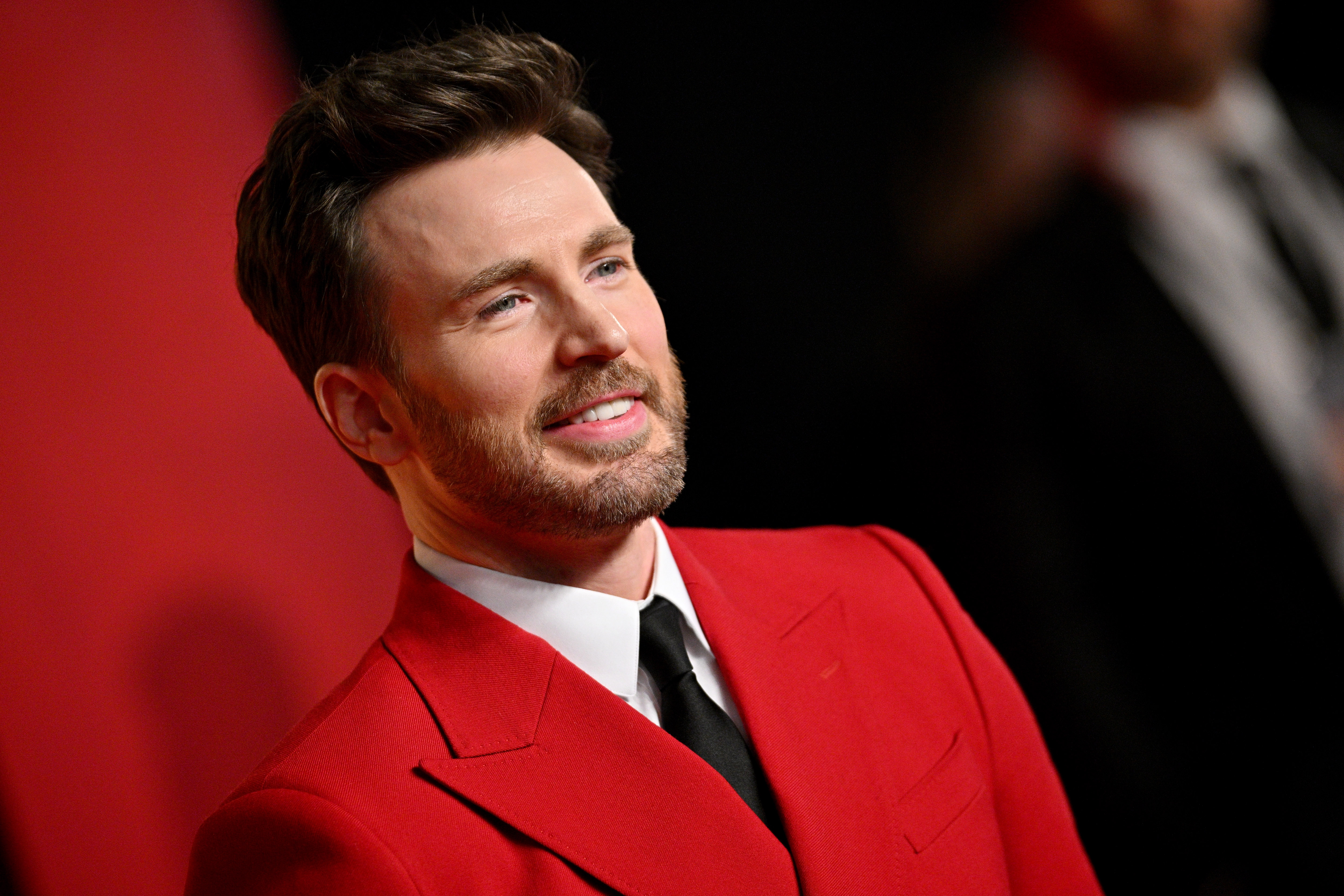 Chris Evans in a red carpet setting, wearing a suit with a tie, smiles while looking slightly away from the camera
