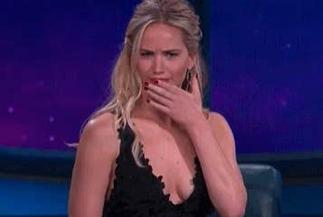 A person with long hair and a perplexed expression covers their mouth while seated, wearing a sleeveless, black patterned dress