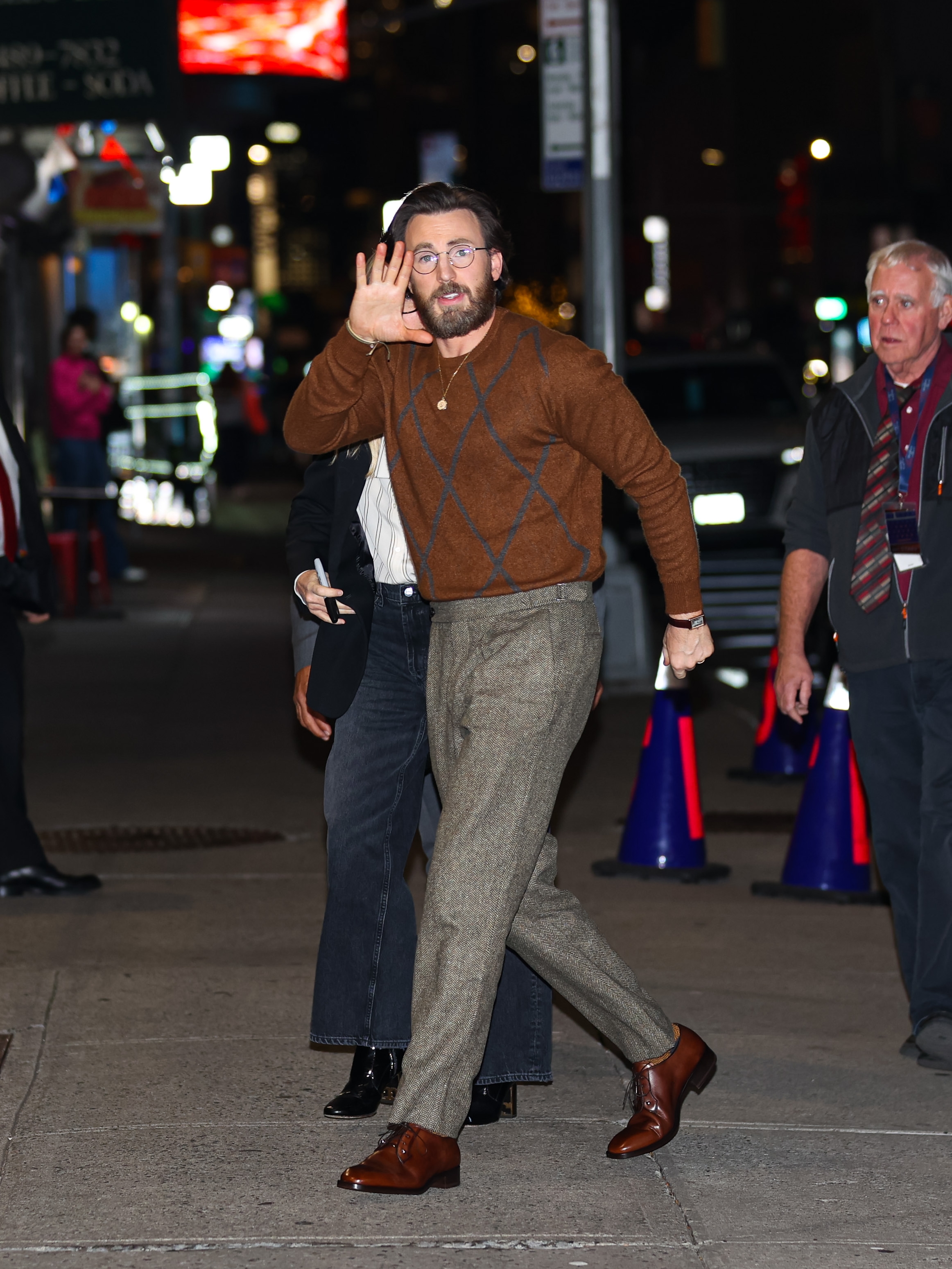 Chris Evans in a sweater and trousers waves on a city street at night, with others around him