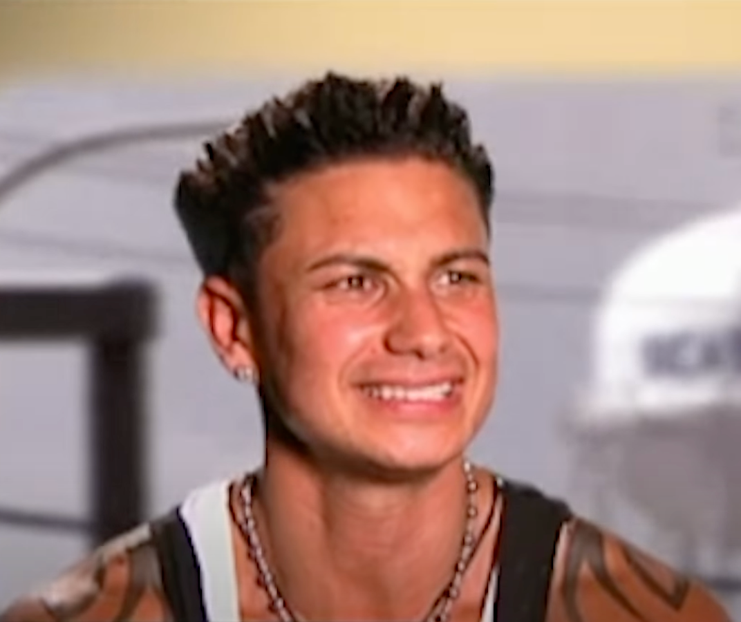 Person with a spiky hairstyle, wearing a necklace and sleeveless top, smiles slightly. The background appears indoors with abstract elements