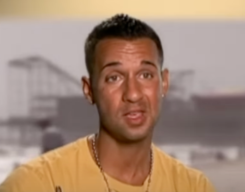 Person speaking to the camera in a casual setting, wearing a simple t-shirt and chain necklace. Background is blurred