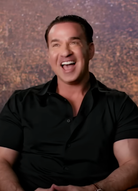 Man in a short-sleeved black shirt laughing, seated against a textured background