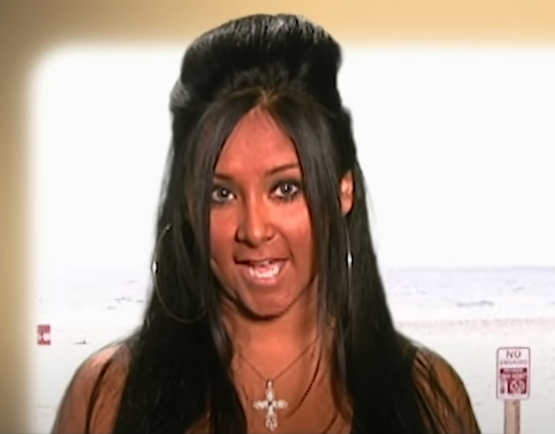 Person with long black hair styled in a bump, wearing hoop earrings and a cross necklace, speaking on a beach background, possibly in a TV show