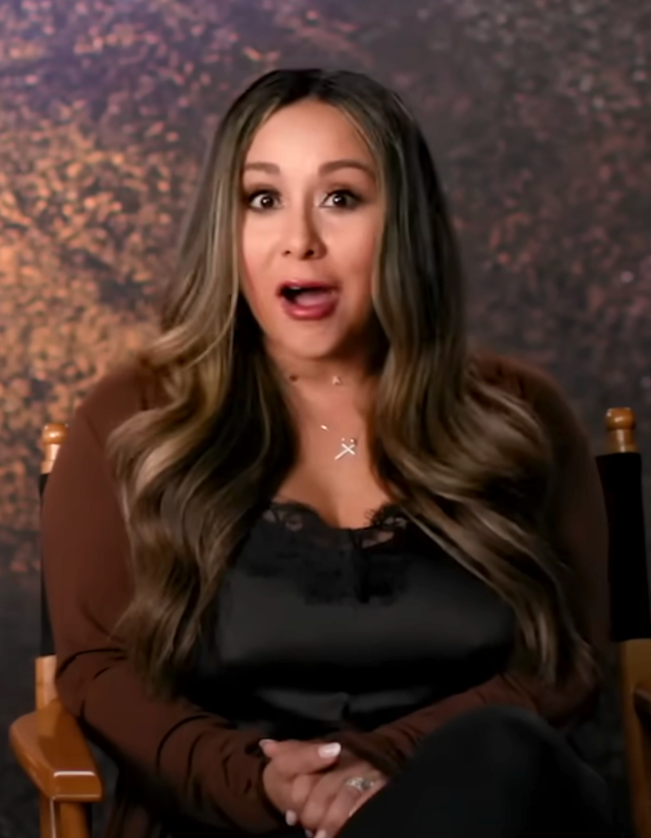 A person with long hair sits in a director's chair, wearing a lace-trimmed top and open cardigan, looking surprised during an interview