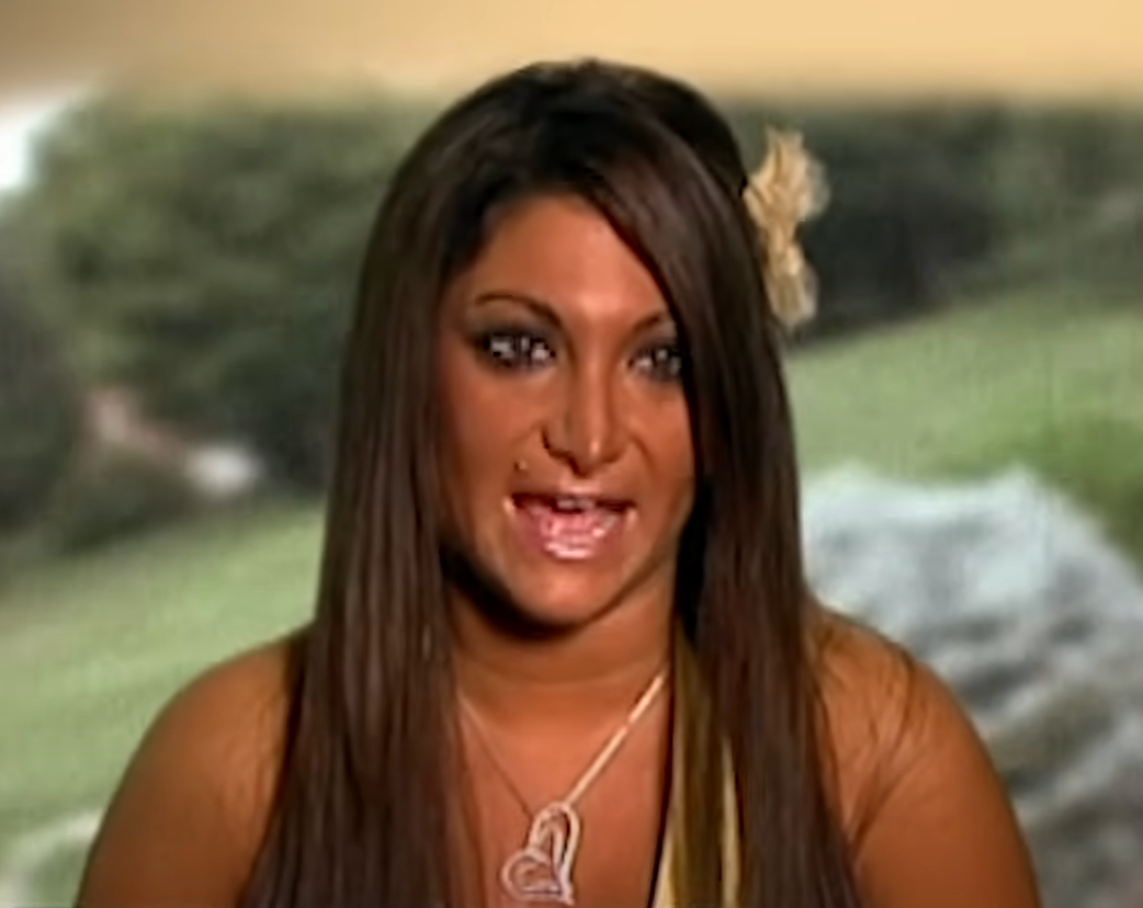 Person with long hair and flower accessory, wearing a necklace. Background features a blurred outdoor scene