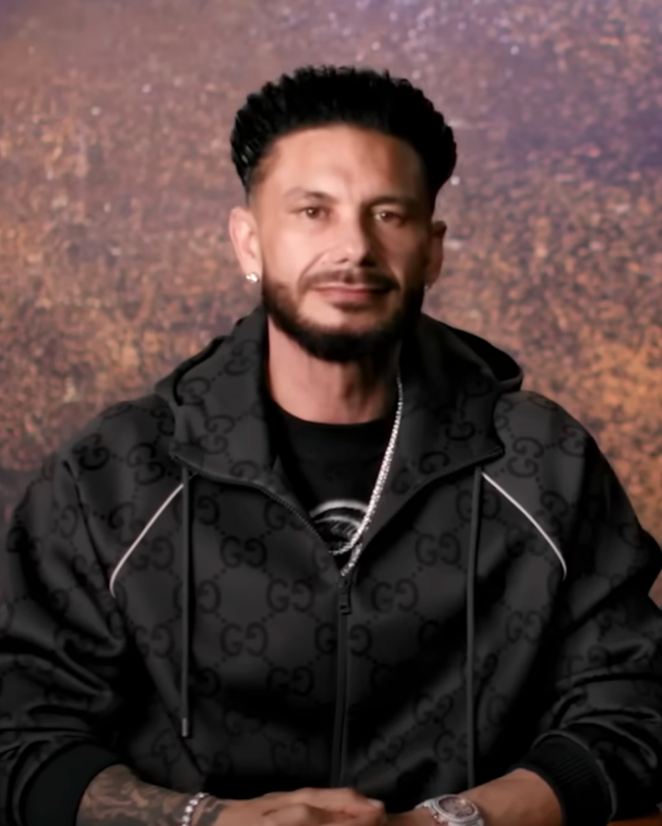 Person wearing a patterned jacket and a chain necklace, sitting against a textured backdrop