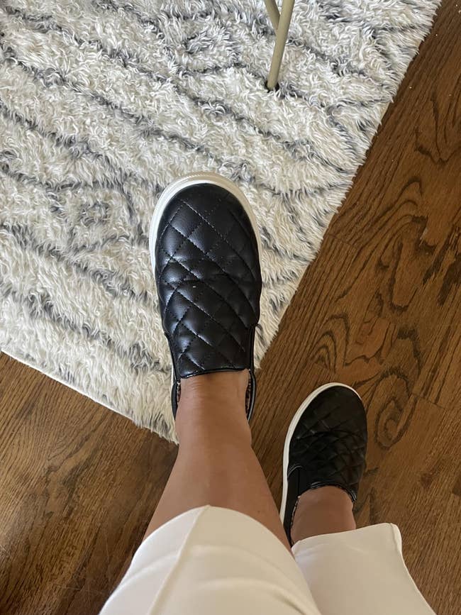 Person wearing quilted slip-on shoes and airy pants stands connected hardwood and patterned rug