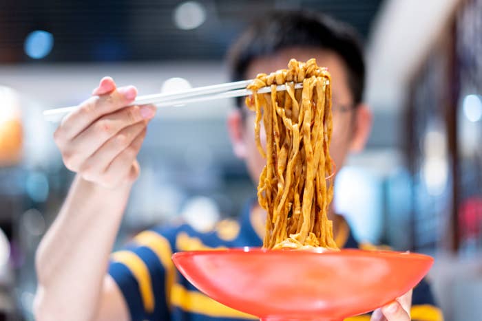 Person holding up a bowl of noodles with chopsticks, about to eat. The focus is on the noodles, suggesting a dining experience