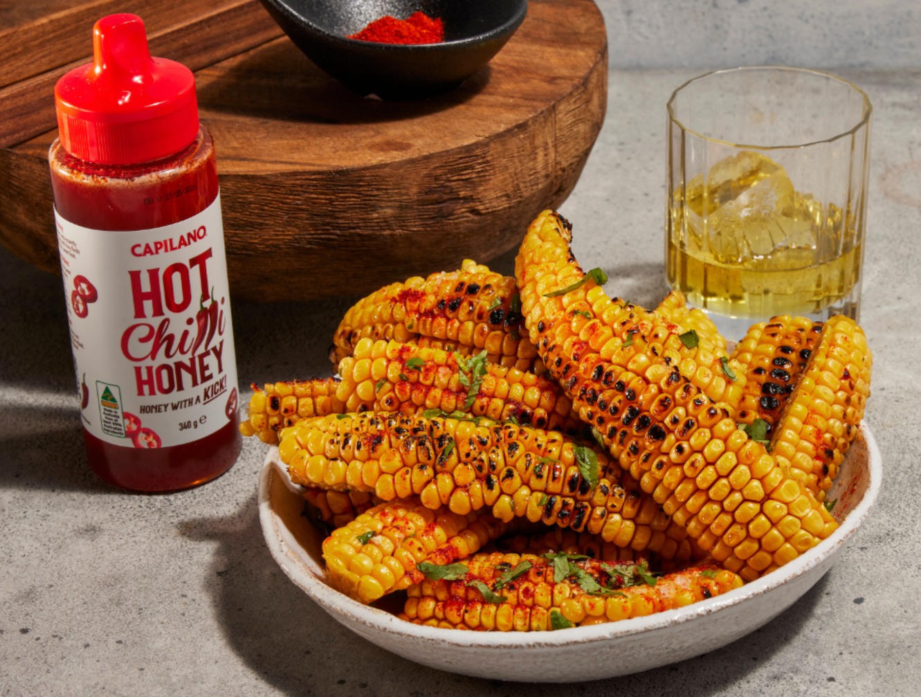 Grilled corn pieces drizzled with Capilano Hot Chili Honey, next to a glass of beverage and a wooden board with spices