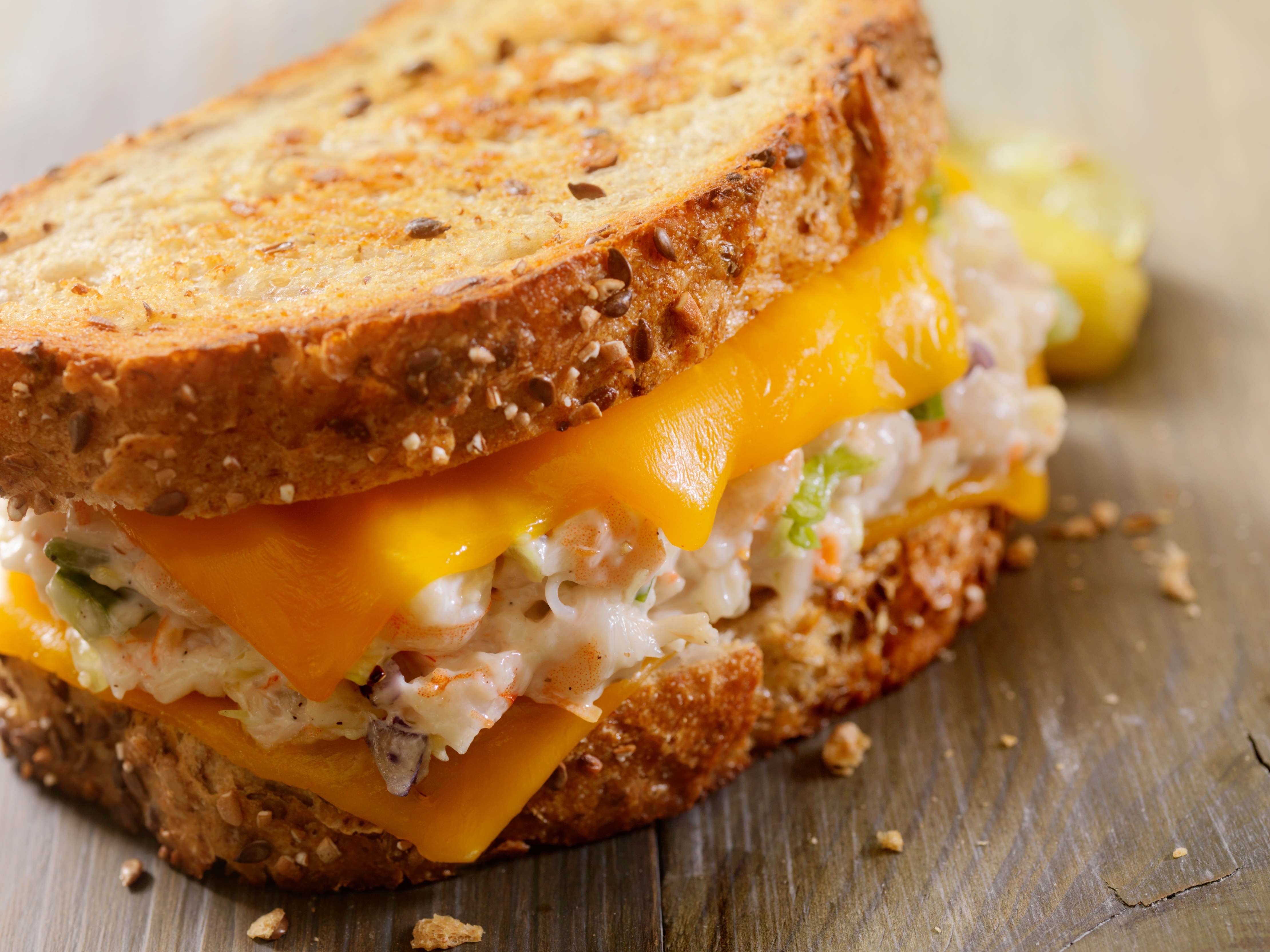 A sandwich with cheddar cheese, creamy seafood salad, and vegetables, served on toasted whole-grain bread, sits on a wooden surface