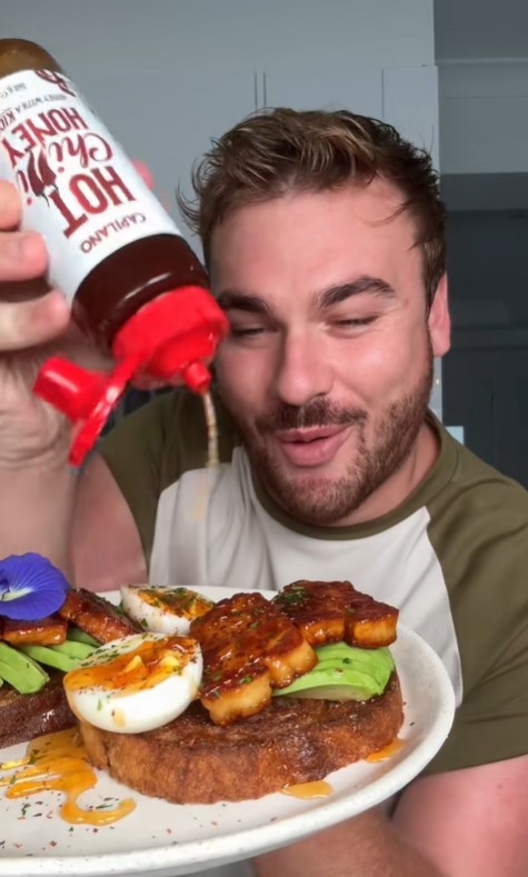 Man smiling and drizzling hot honey over toast with avocado, eggs, and glazed chunks