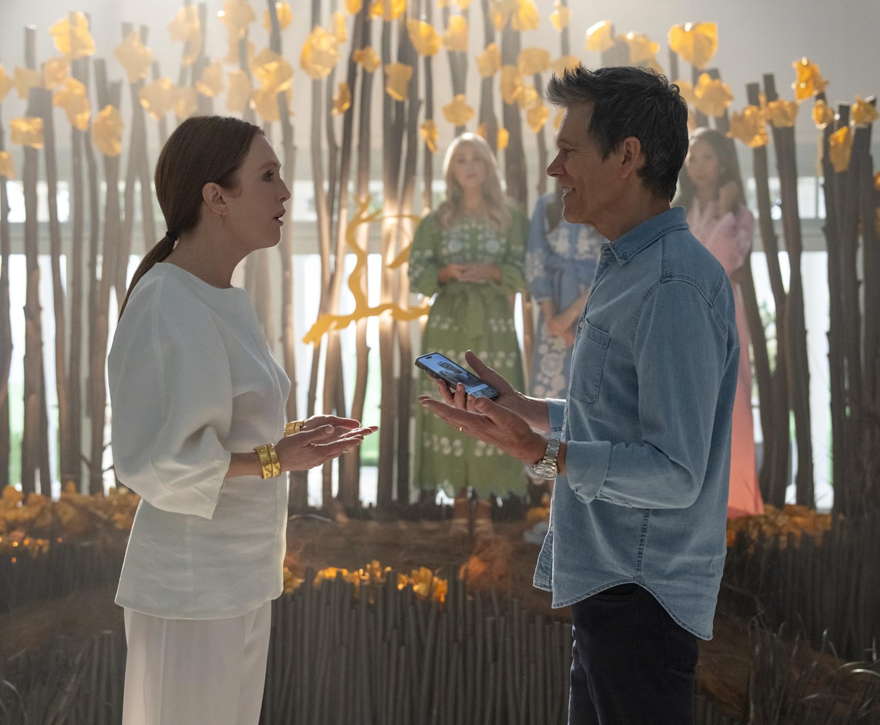 Two people in conversation, one wearing a white outfit and gold bracelet, the other in a denim shirt. Background features others and decorative sticks